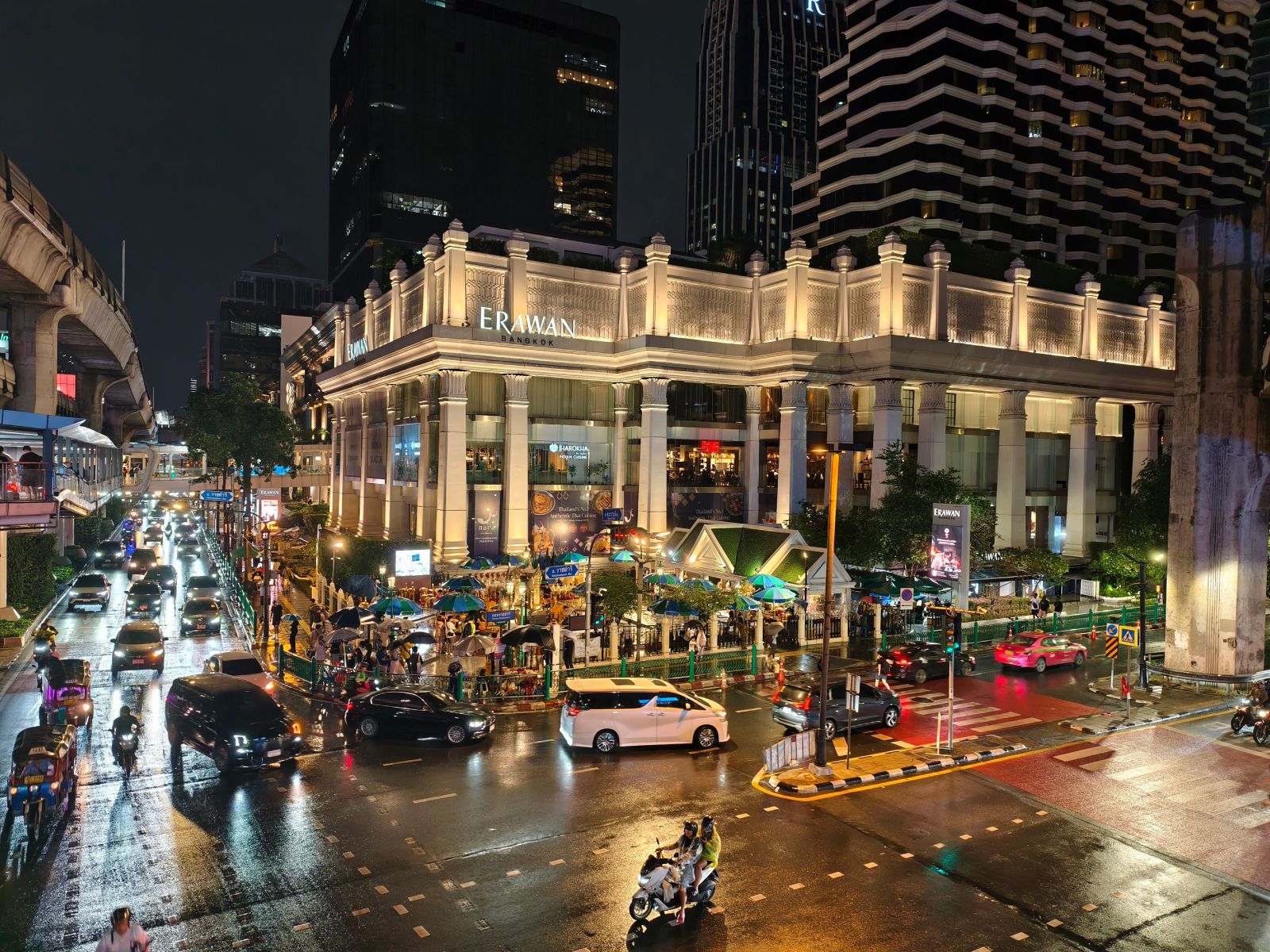 The bustling street scene at night near Erawan, Bangkok, showcases urban life, traffic, and illuminated architecture.